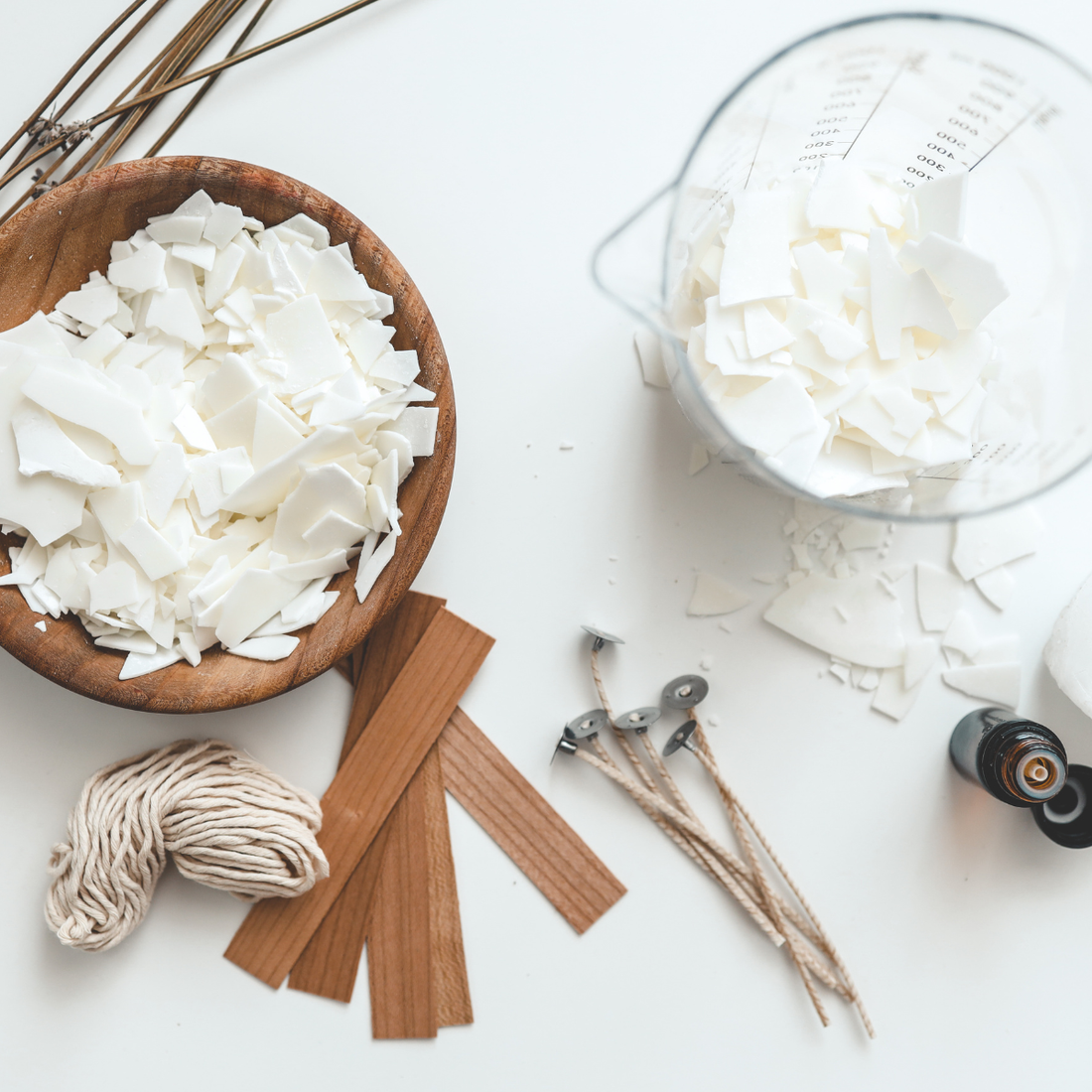 Sage & Jade Co. a flat lay photo of soy wax in a wooden bowl and measuring cup, wooden wicks, cotton wicks, cotton twine, a fragrance oil bottle.